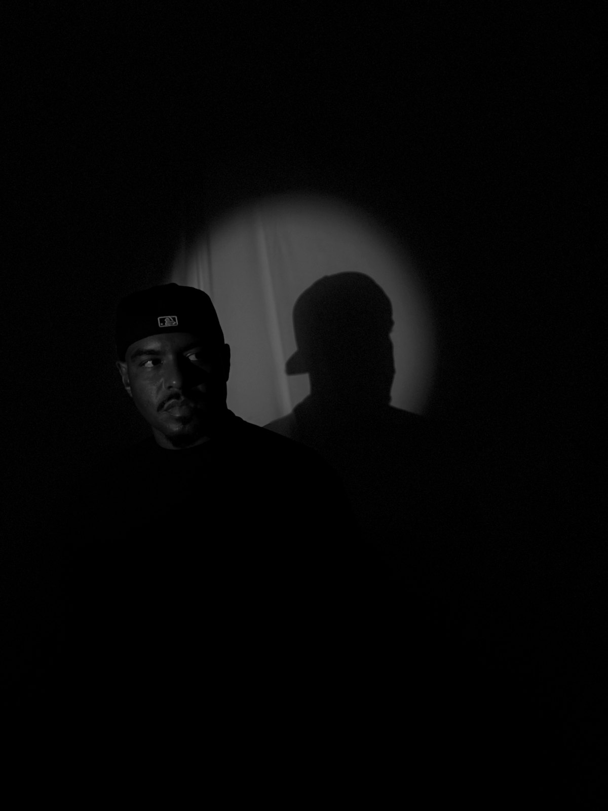Retrato em preto e branco de Fabrício Crepaldi, de boné, emergindo do escuro, com a própria sombra projetada dentro de um círculo de luz na parede.